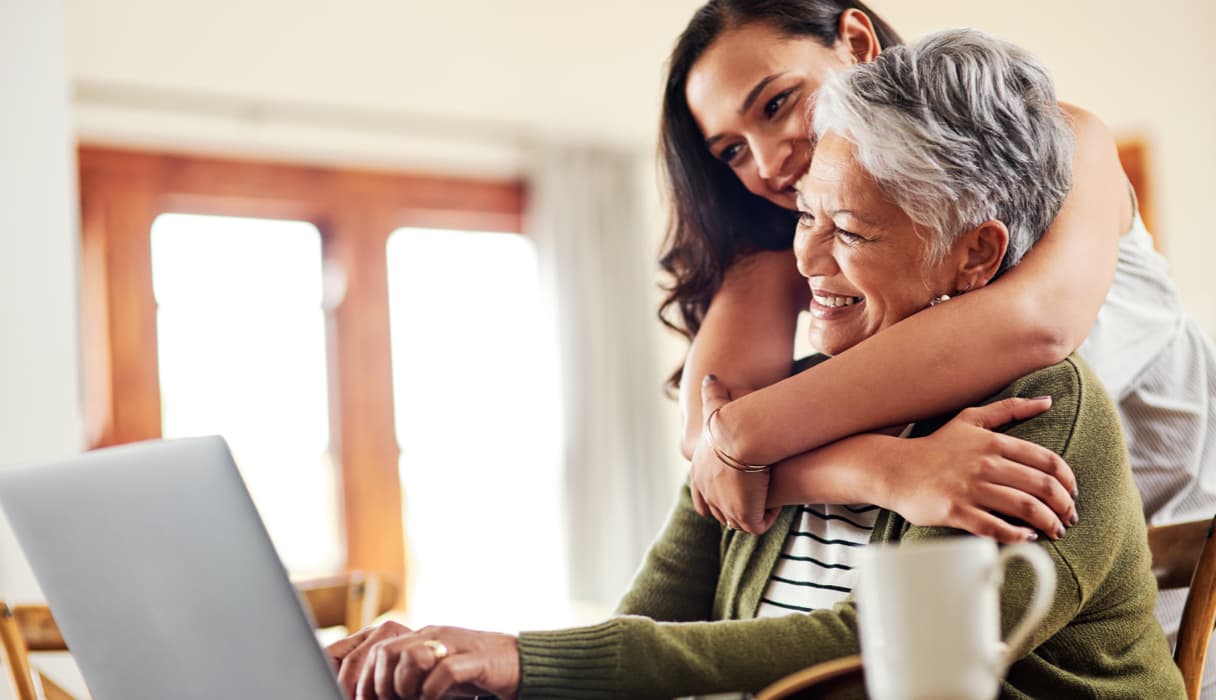 A woman hugging a grandmother figure while they both smile and look forward at a laptop in a home like setting.